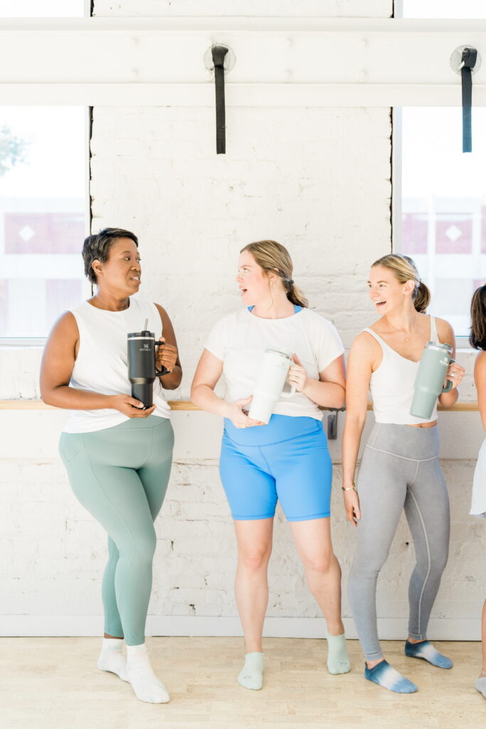 Three women smiling together while holding yoga mats at a fitness studio